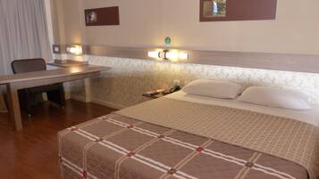 Quarto de hotel com cama, escrivaninha e cadeira. Há luminárias na parede acima da escrivaninha e uma mesinha de cabeceira com abajur.