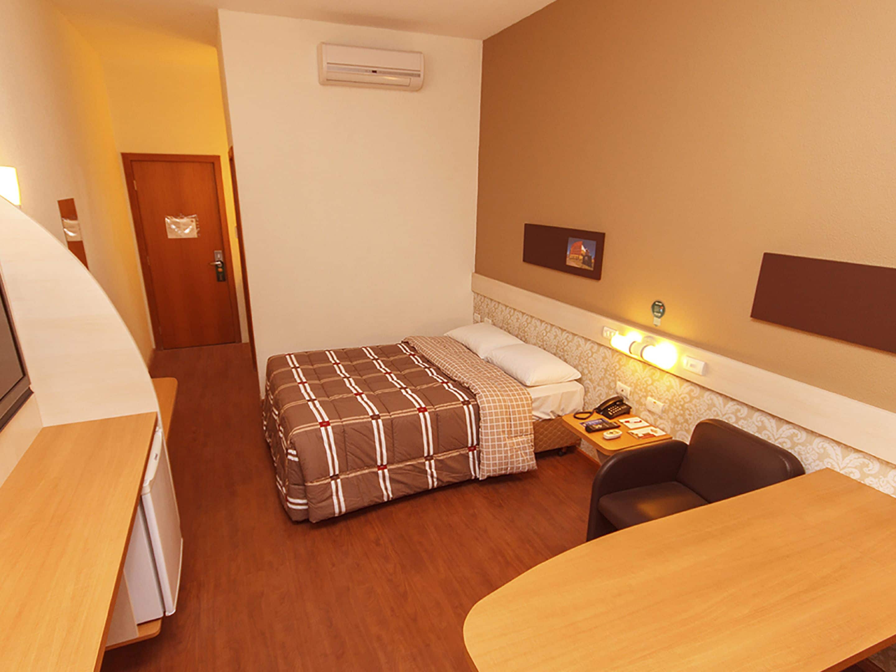 A hotel room with a bed, a desk, a chair, a bedside table with a phone, and an air conditioning unit on the wall.