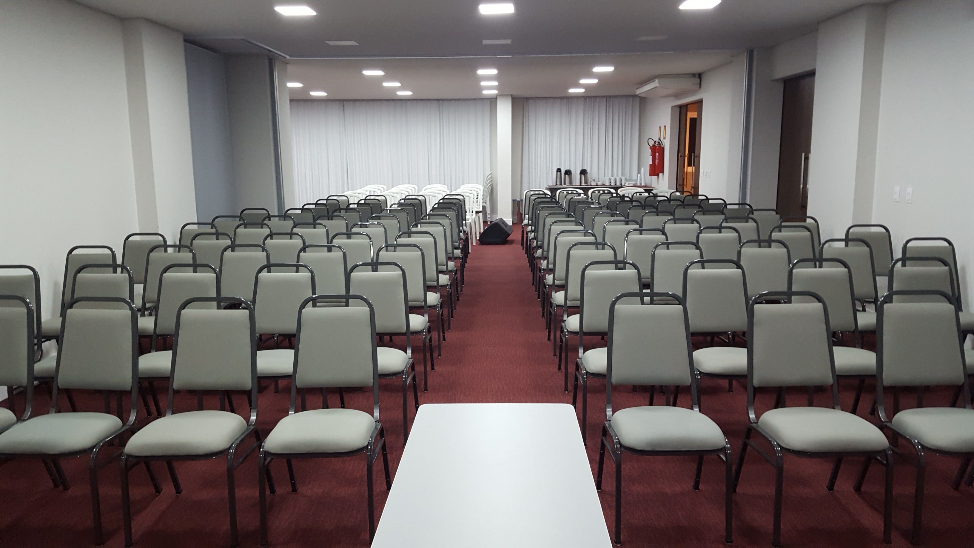 A conference room with rows of chairs and a long table, white walls, and ceiling lights.