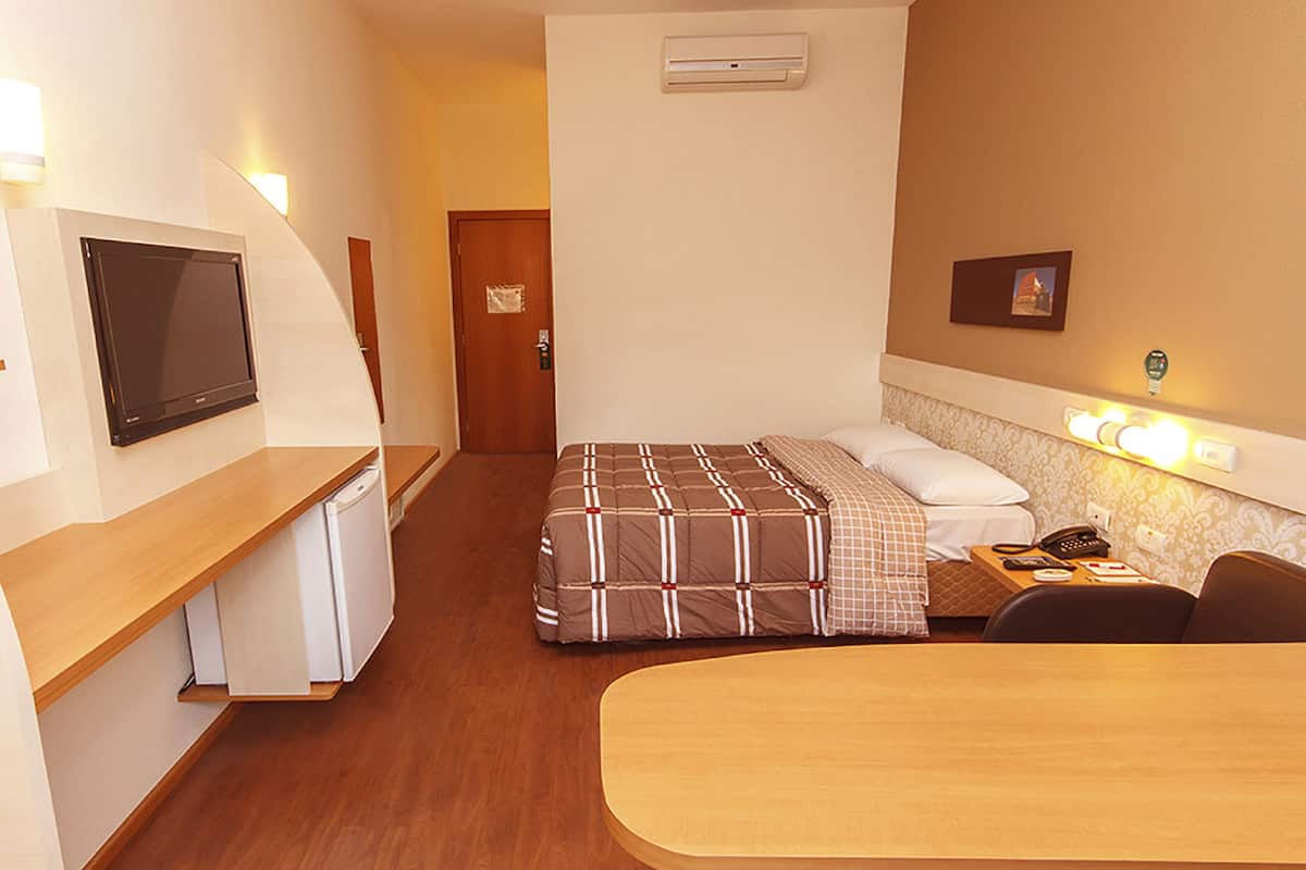 Habitación de hotel compacta con cama, escritorio, televisor y una pequeña kitchenette.