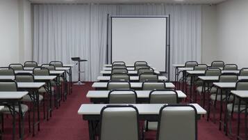 Un salón de conferencias con filas de mesas y sillas, una pantalla de proyector y un fondo de cortina blanca.