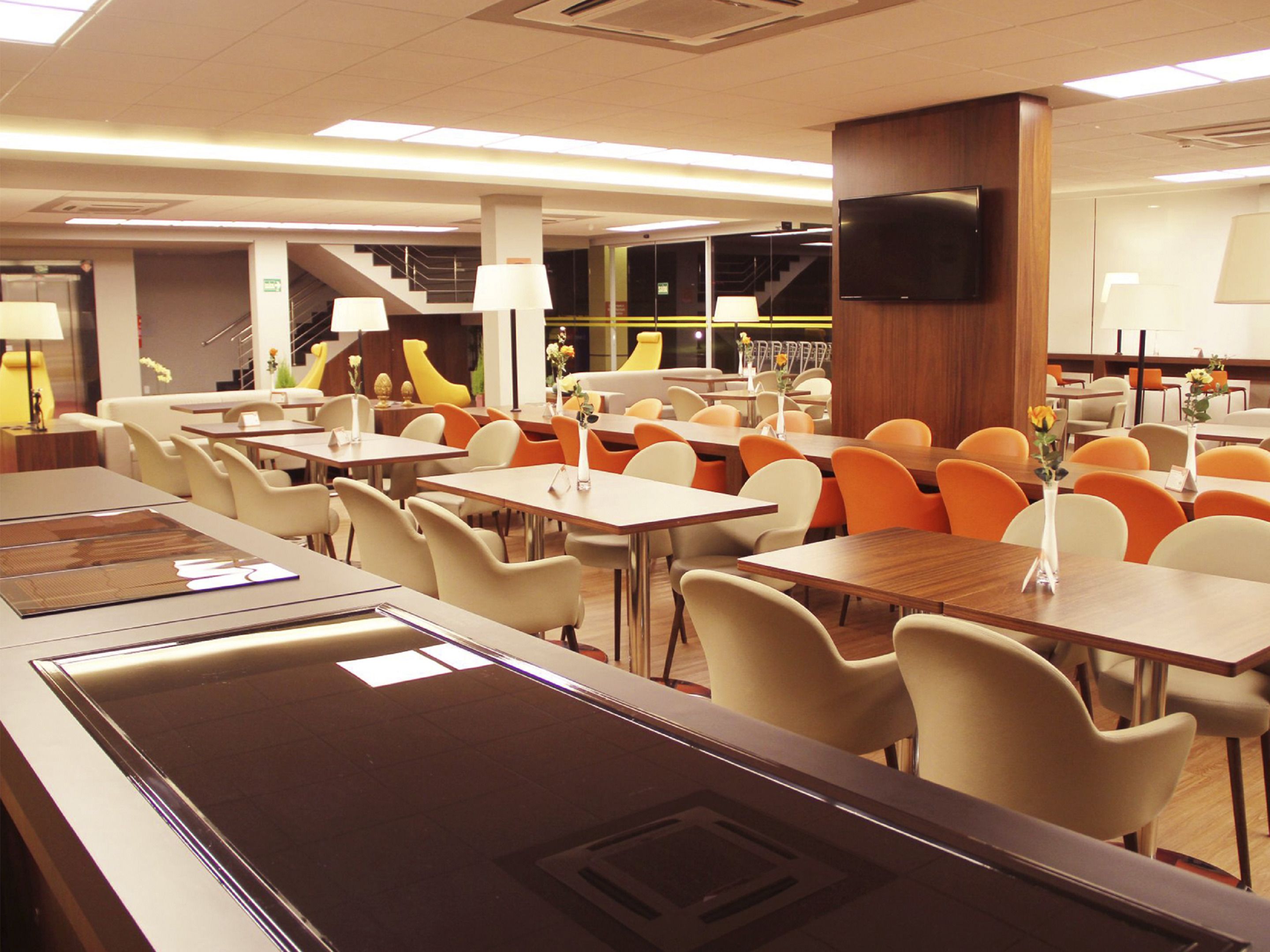 A modern dining area with multiple tables and chairs, a central staircase, and a flat-screen TV mounted on the wall.