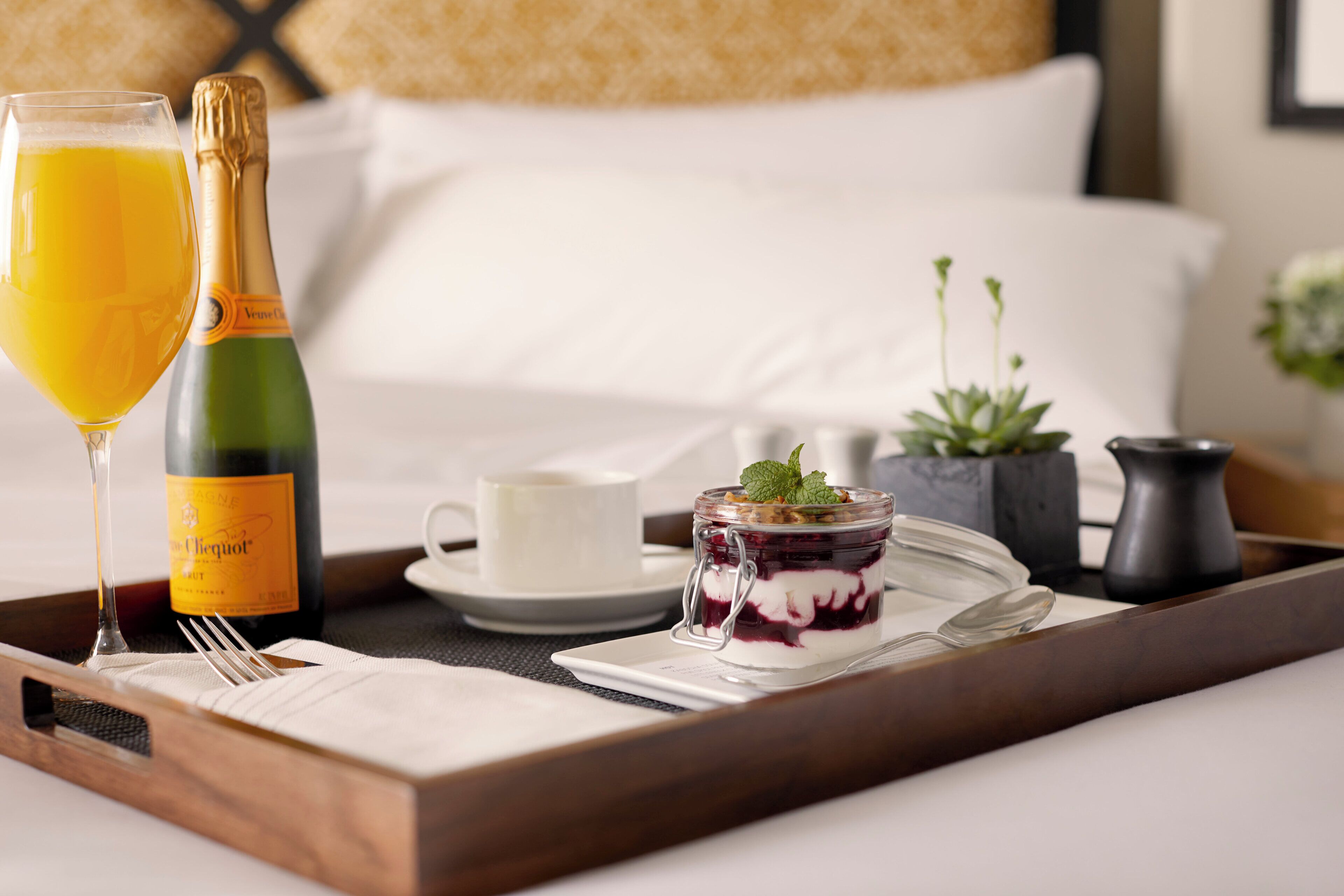 En bakke med morgenmad, inklusive et glas appelsinjuice, en champagneflaske, en lille dåse dessert, en kop og en ske på en træbakke.