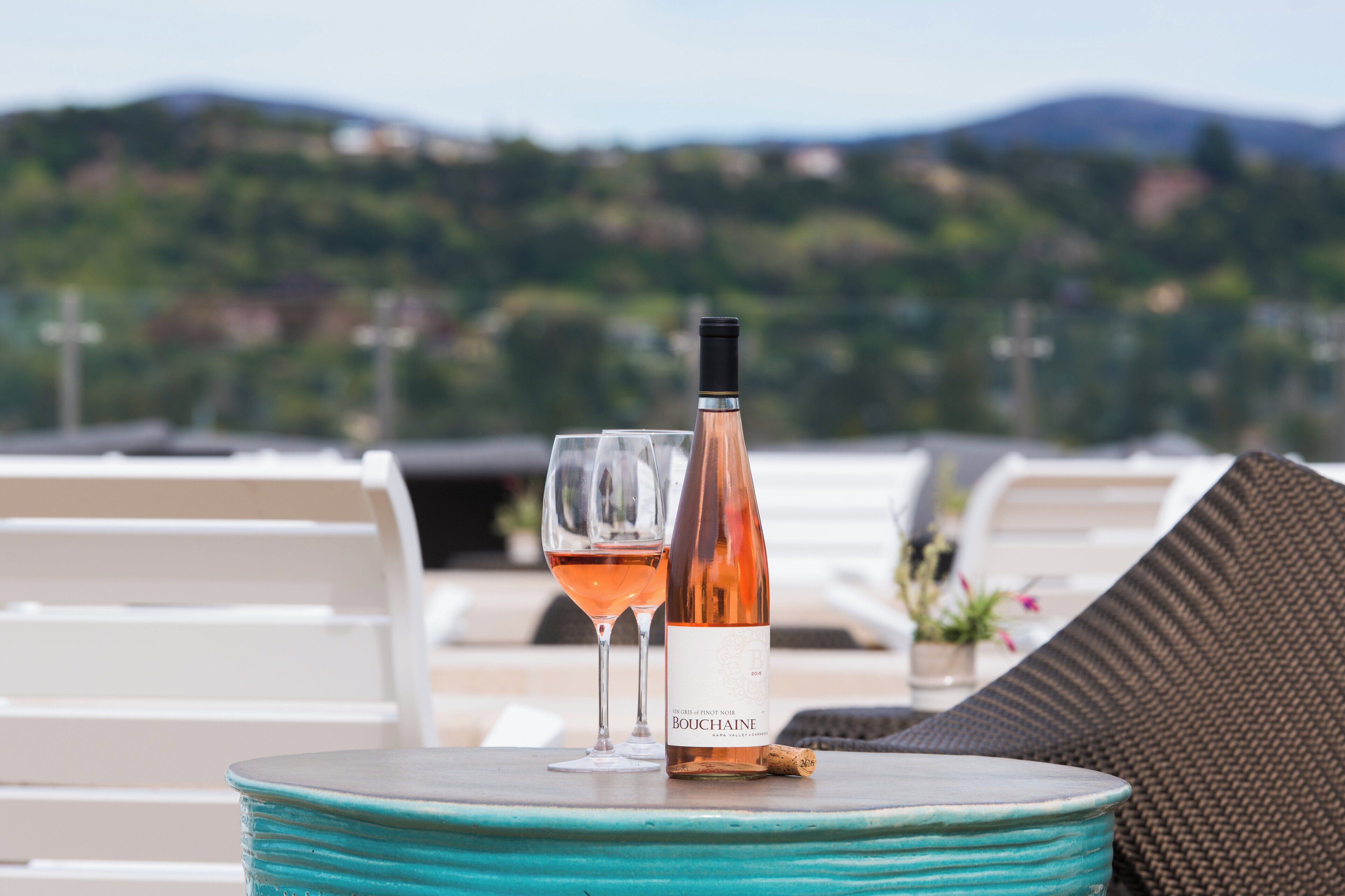 A table with a bottle of Bouchanée wine and two glasses on a turquoise table, set against a backdrop of white lounge chairs and a mountainous landscape.
