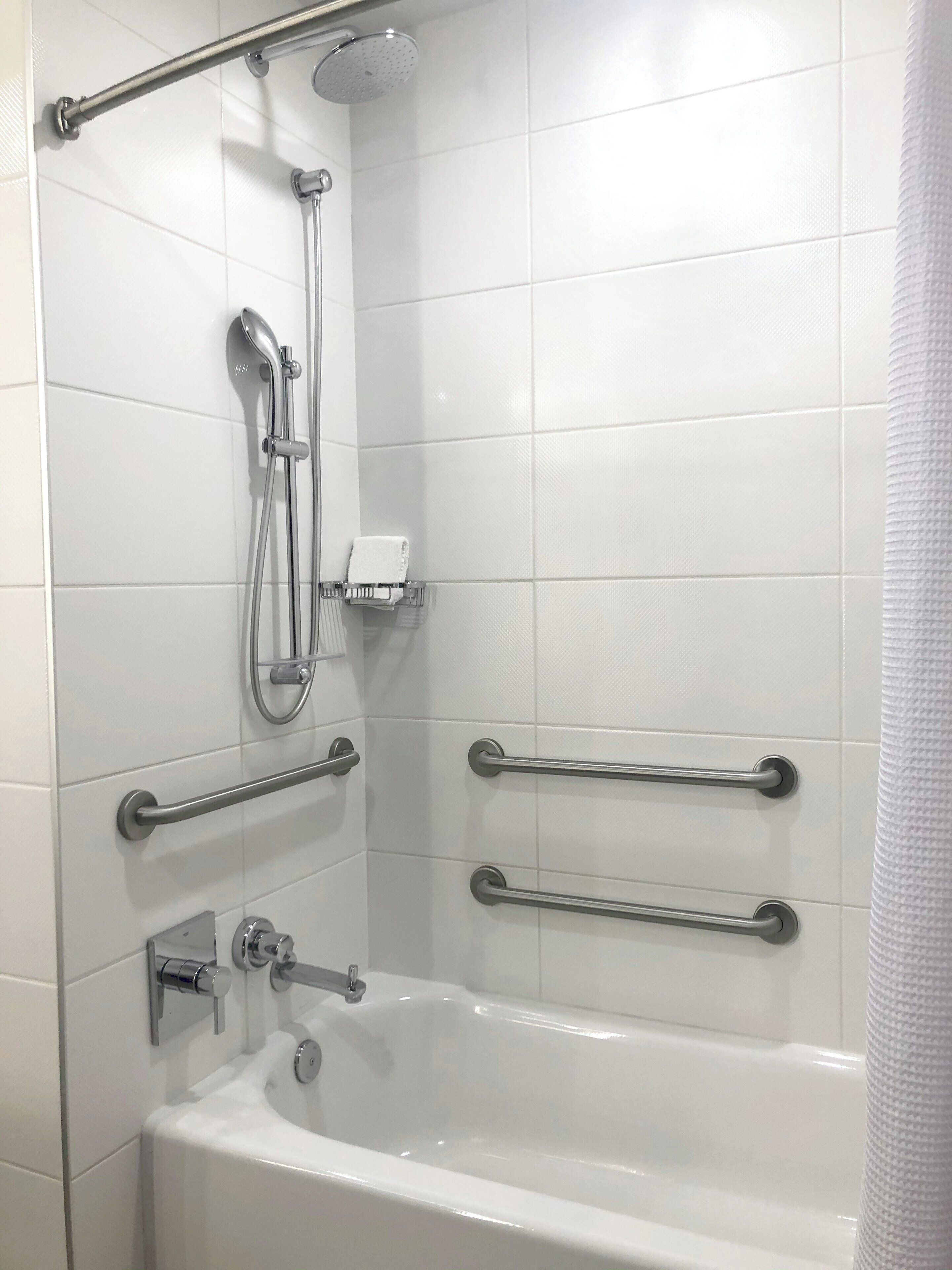 Casa de banho moderna com azulejos brancos, banheira, chuveiro com chuveiro manual e barras de apoio para segurança.