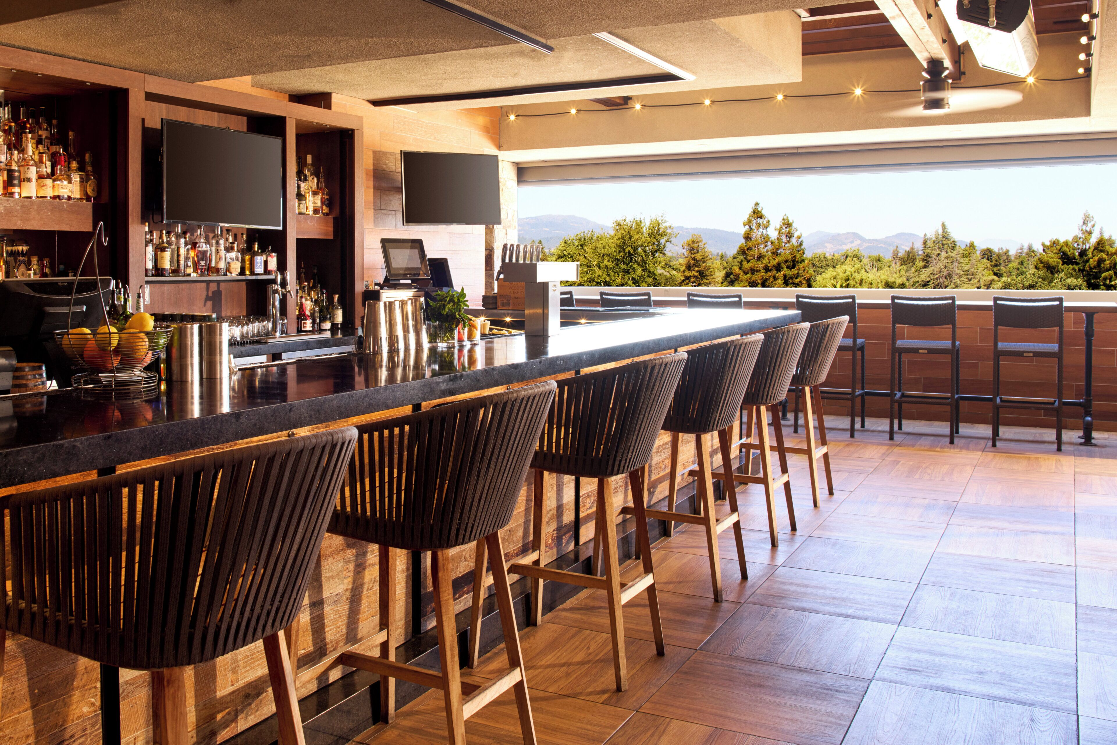 Zona de bar con una barra alargada, varios taburetes, un televisor y vistas al exterior.