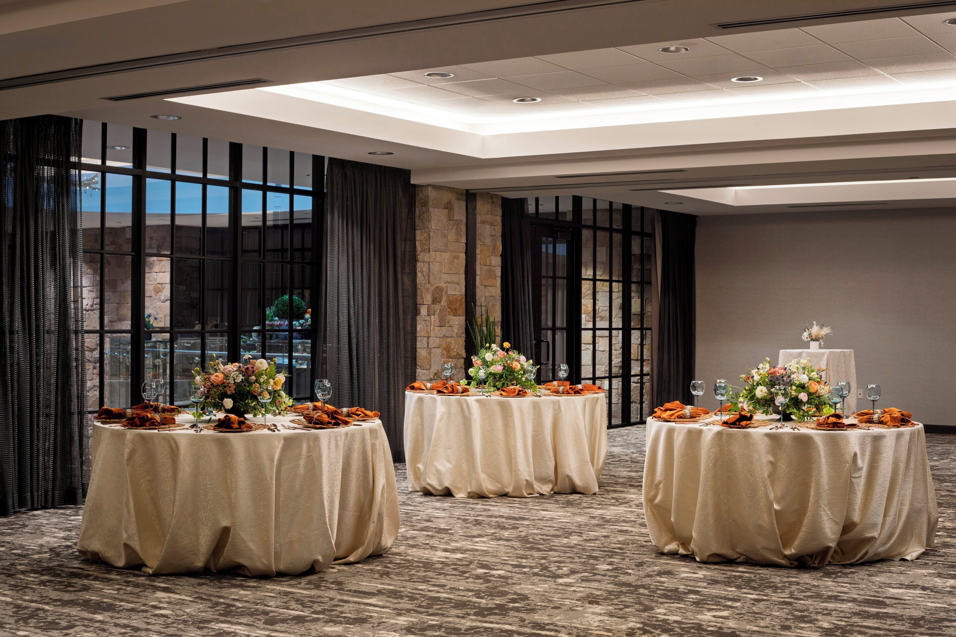 A conference room with three round tables, each covered with a beige tablecloth and set with orange napkins, wine glasses, and floral centerpieces.