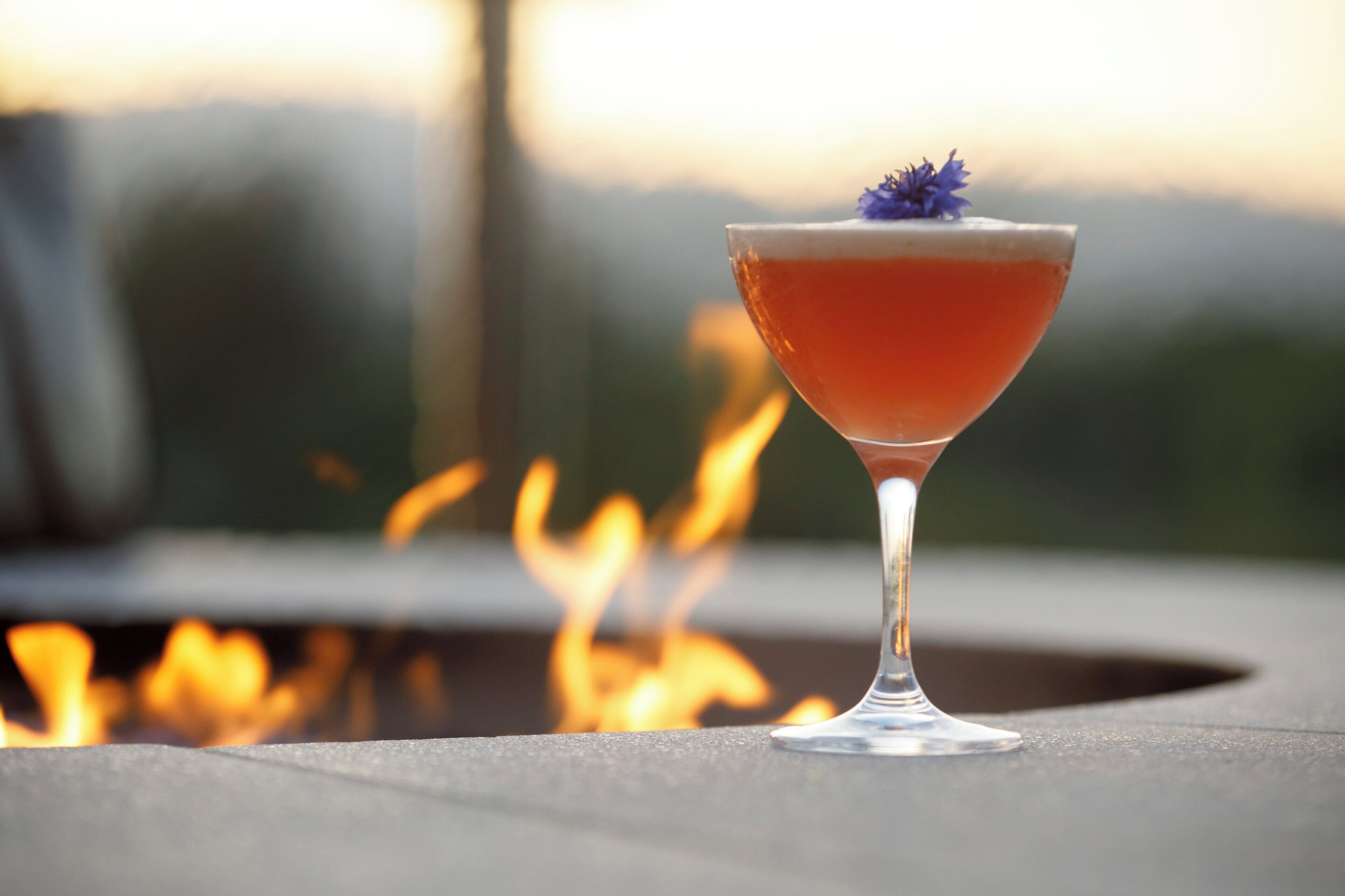 A cocktail in a martini glass with a garnish, placed on a table with a blurred background of fire.