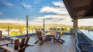 An outdoor seating area with wooden tables and chairs, a fire pit, and a view of the city and mountains. - Archer Hotel Napa (Napa)