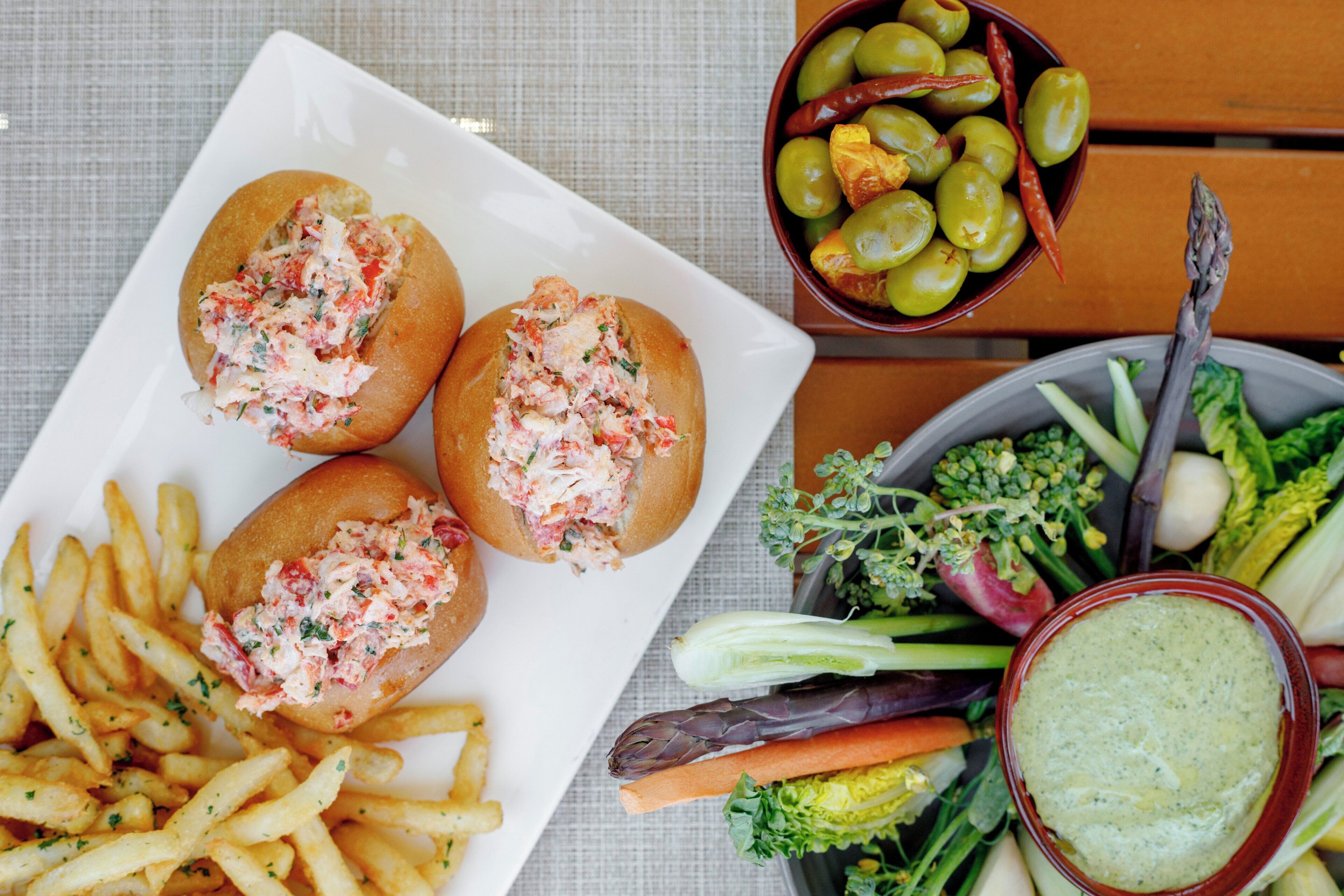 A plate with lobster rolls, a bowl of olives, and a plate of vegetables.