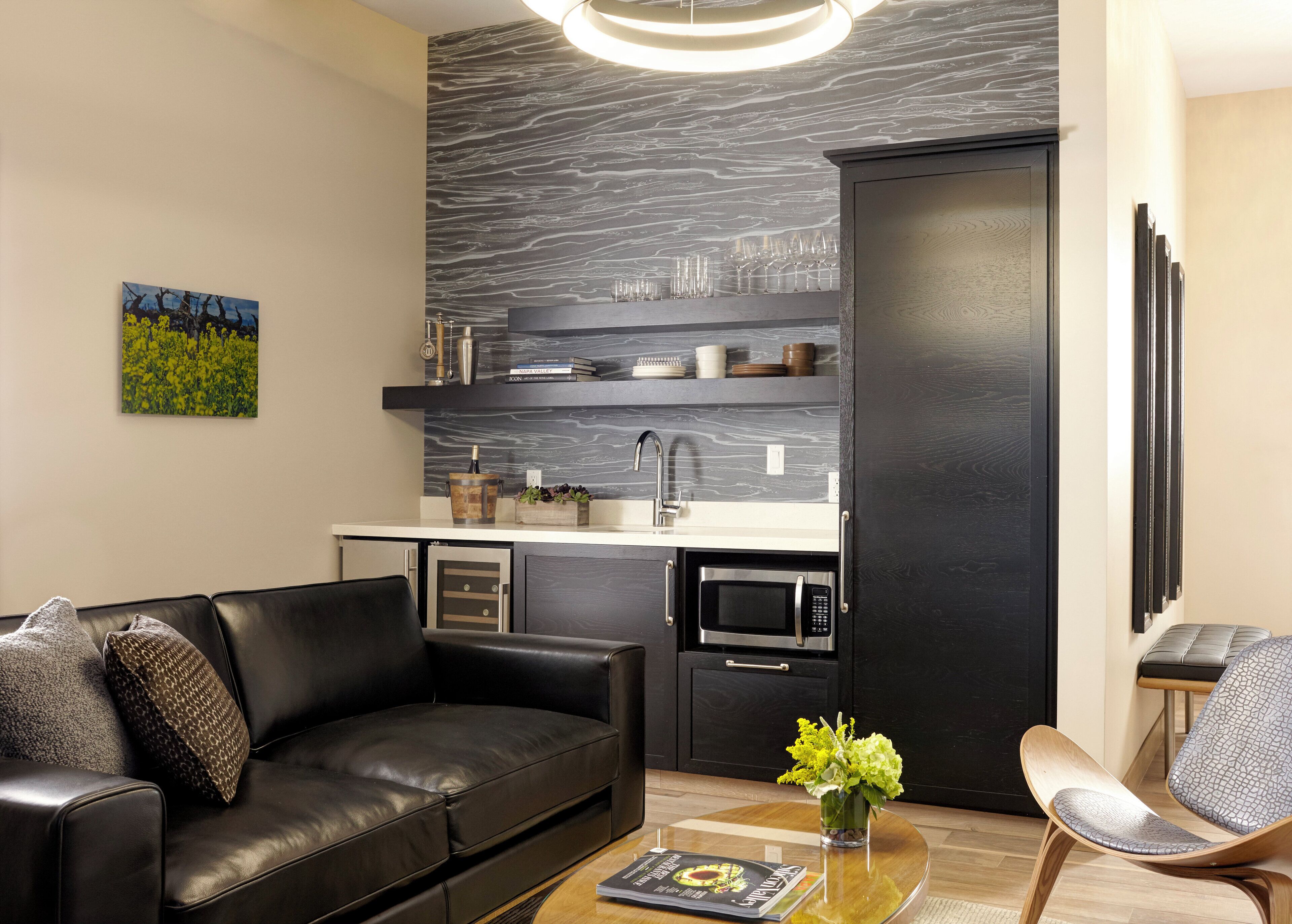 Una sala de estar moderna con un sofá de cuero negro, una mesa de centro de madera con una revista, un jarrón pequeño con flores, y una zona de cocina con muebles oscuros, un microondas y una botella de vino.