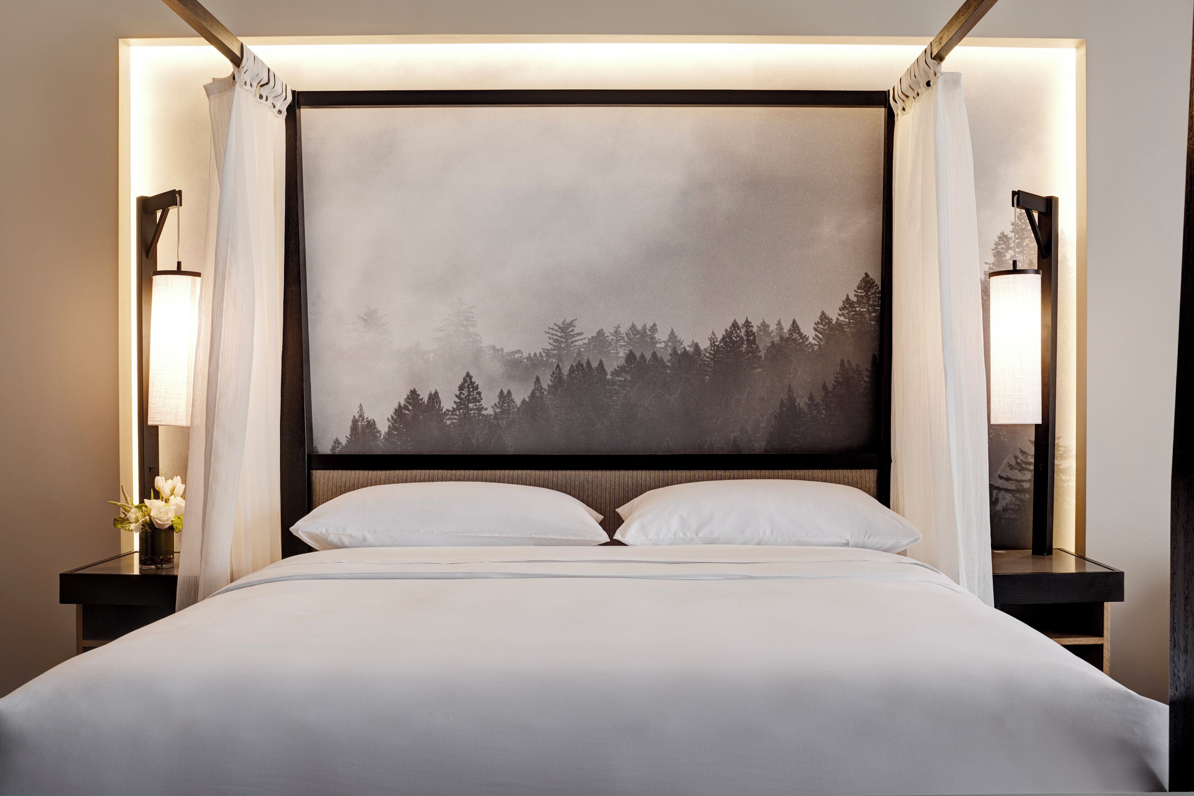Een keurig opgemaakt bed met daarboven een groot, ingelijst plaatje van een bebost landschap.
