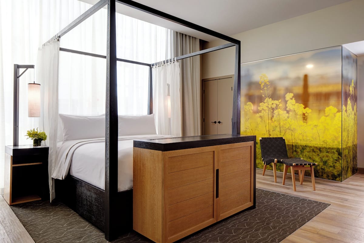 A four-poster bed with a wooden cabinet, a chair, and a large window with curtains.
