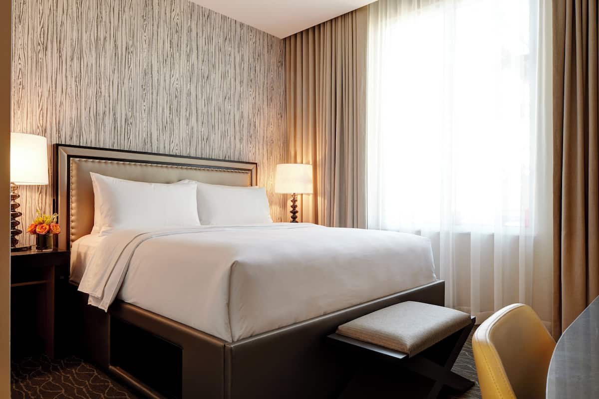A hotel room with a large bed, two bedside tables with lamps, a bench at the foot of the bed, and a view of the outdoors through sheer curtains.
