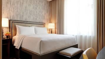 Quarto de hotel com uma cama grande, duas mesas de cabeceira com luminárias, um banco aos pés da cama e vista para o exterior através de cortinas translúcidas.