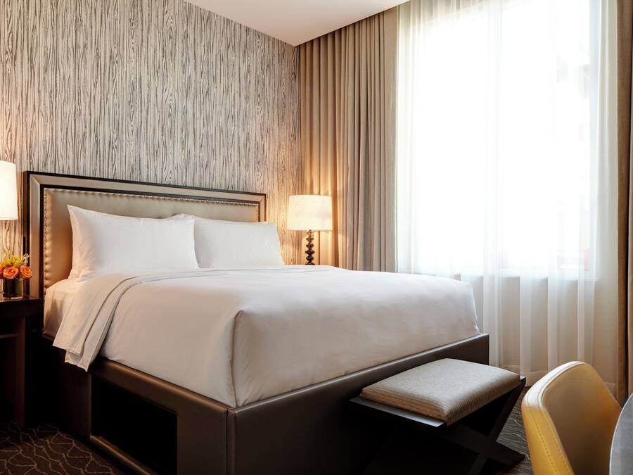 Quarto de hotel com uma cama grande, duas mesas de cabeceira com luminárias, um banco aos pés da cama e vista para o exterior através de cortinas translúcidas.