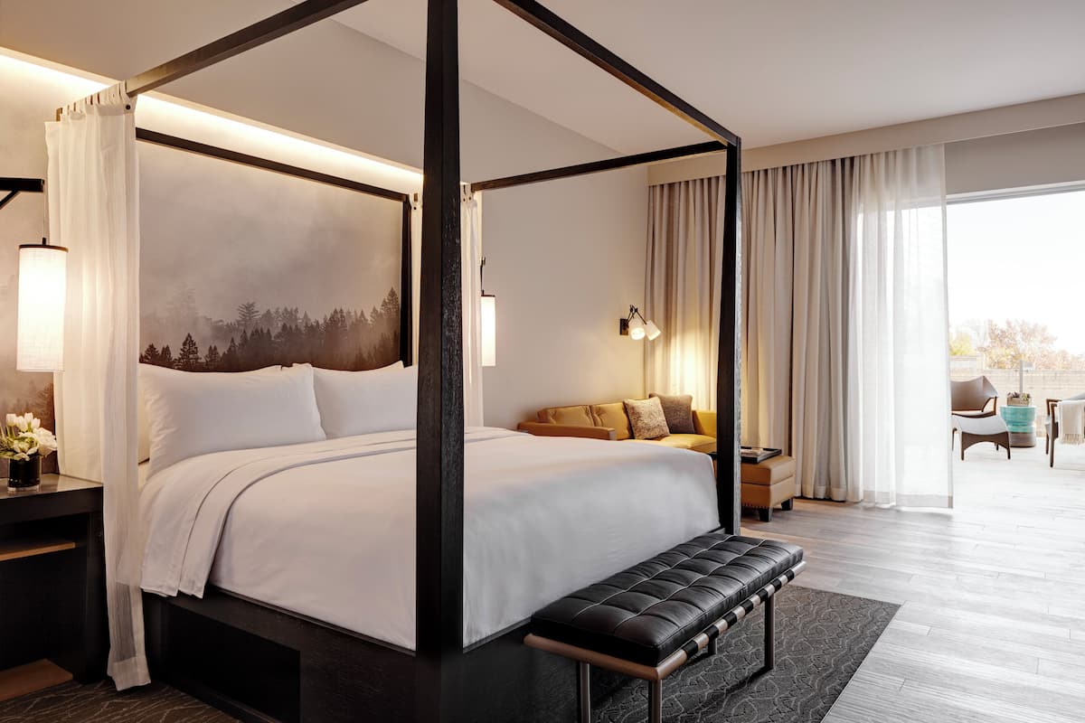 A four-poster bed with a view of a forest through the window, a sofa with cushions, a bench, and a small table with a lamp.