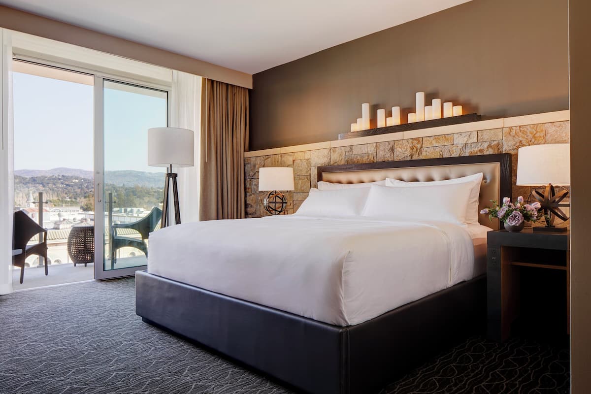 A hotel room with a large bed, a view through a sliding door, a seating area with a chair and table, and a decorative wall with candles.