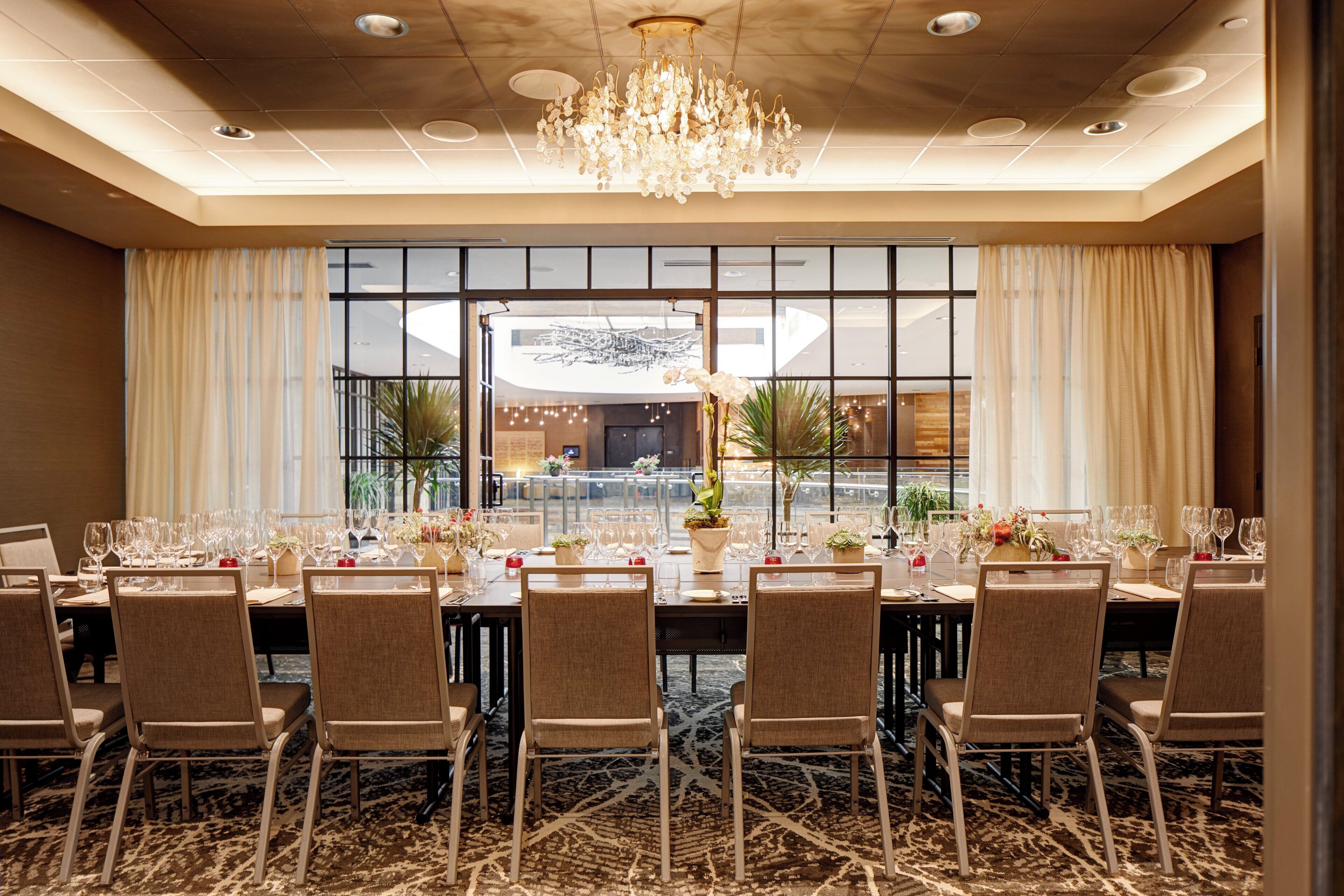 A banquet hall with a long table set for a formal event, surrounded by chairs, with a chandelier and large windows in the background.