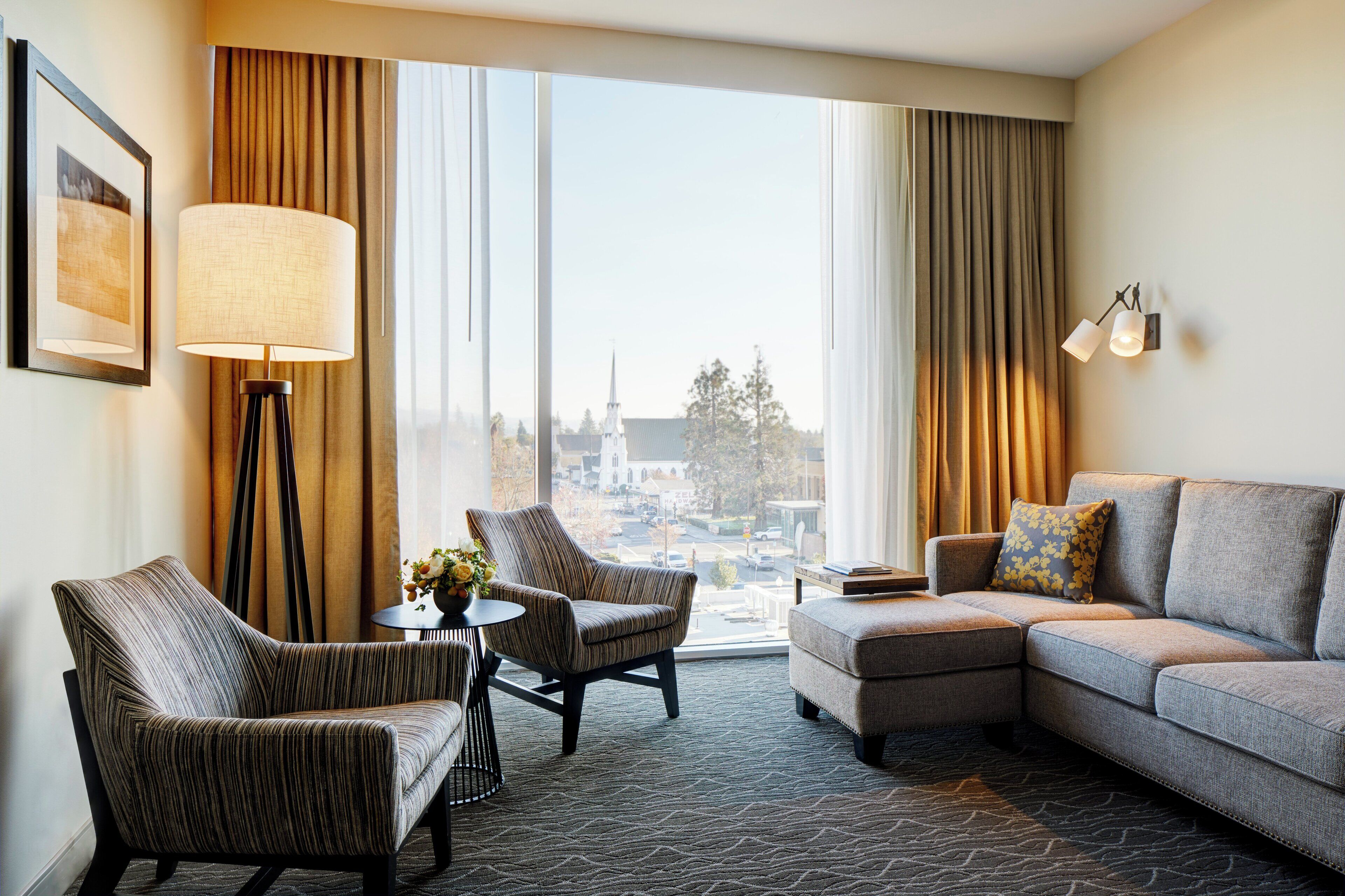 A modern hotel room with a large window, a sofa, two armchairs, a small table with a vase of flowers, and a floor lamp.