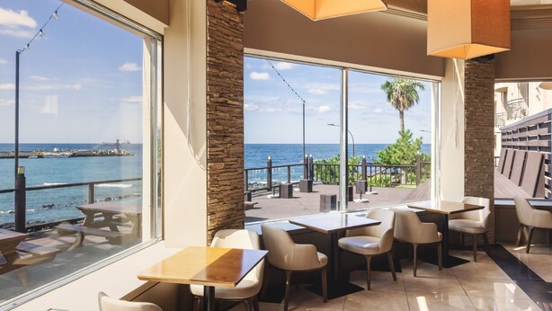 An indoor dining area with large windows overlooking a coastal view, stone accents, and modern lighting fixtures. - Kensington Resort Jeju Hanlim (Jeju City)