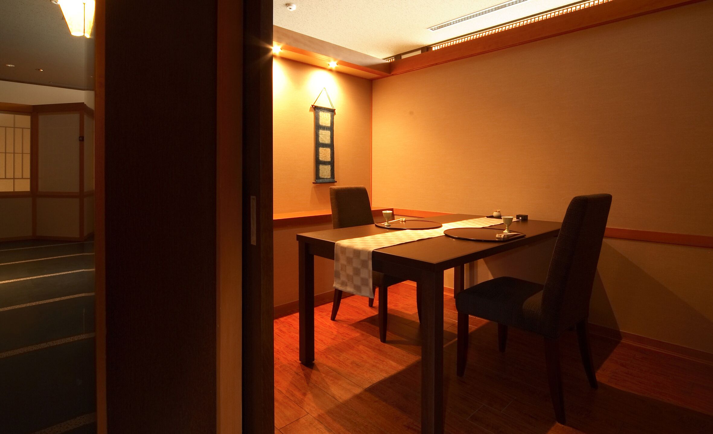 A dining area with a table set for two, a chair, and a wall-mounted light fixture.