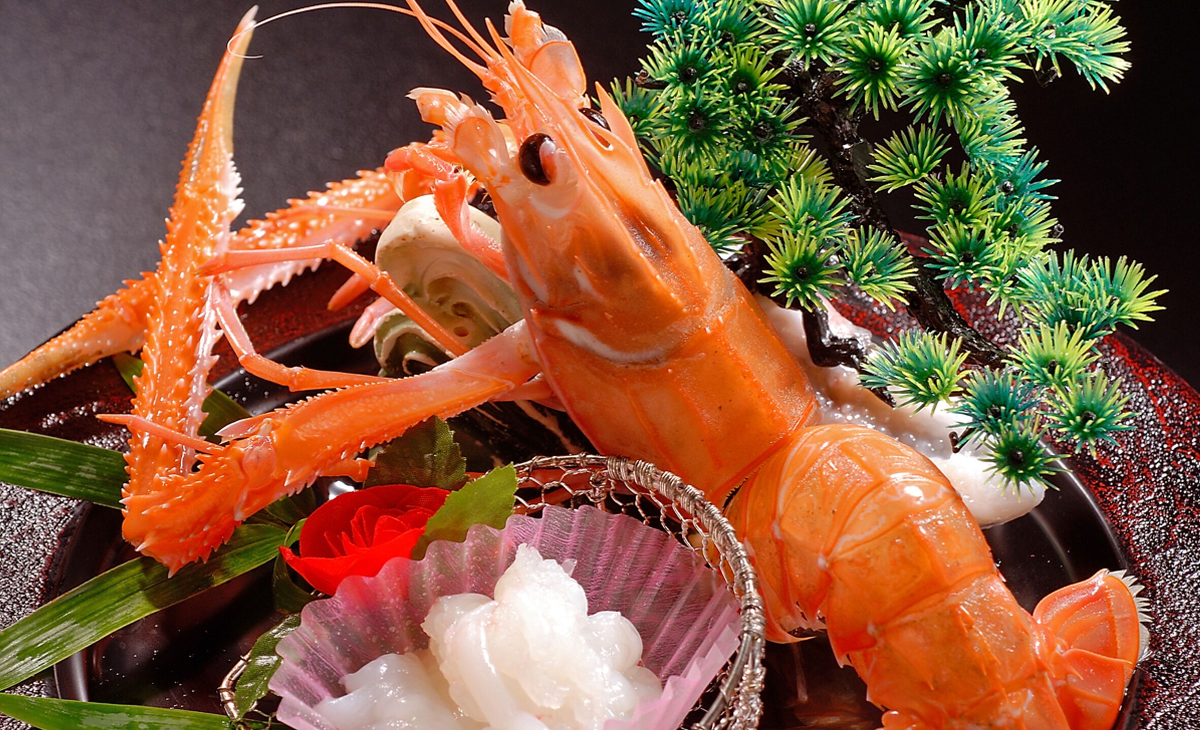 A lobster with a decorative shell and a small bowl of white substance, possibly a sauce, with green foliage in the background.