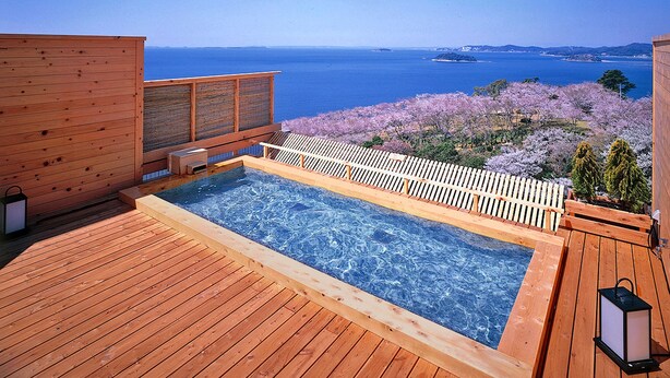 Une terrasse en bois avec un jacuzzi donnant sur un plan d’eau et des arbres en fleurs.