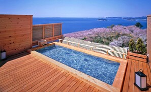 Un patio in legno con vasca idromassaggio che si affaccia su un lago e alberi di ciliegio in fiore.