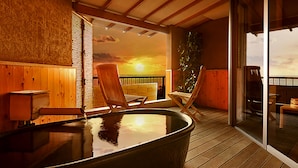 Une terrasse en bois avec un jacuzzi, deux chaises longues et une vue sur le coucher du soleil, visible à travers une porte vitrée.