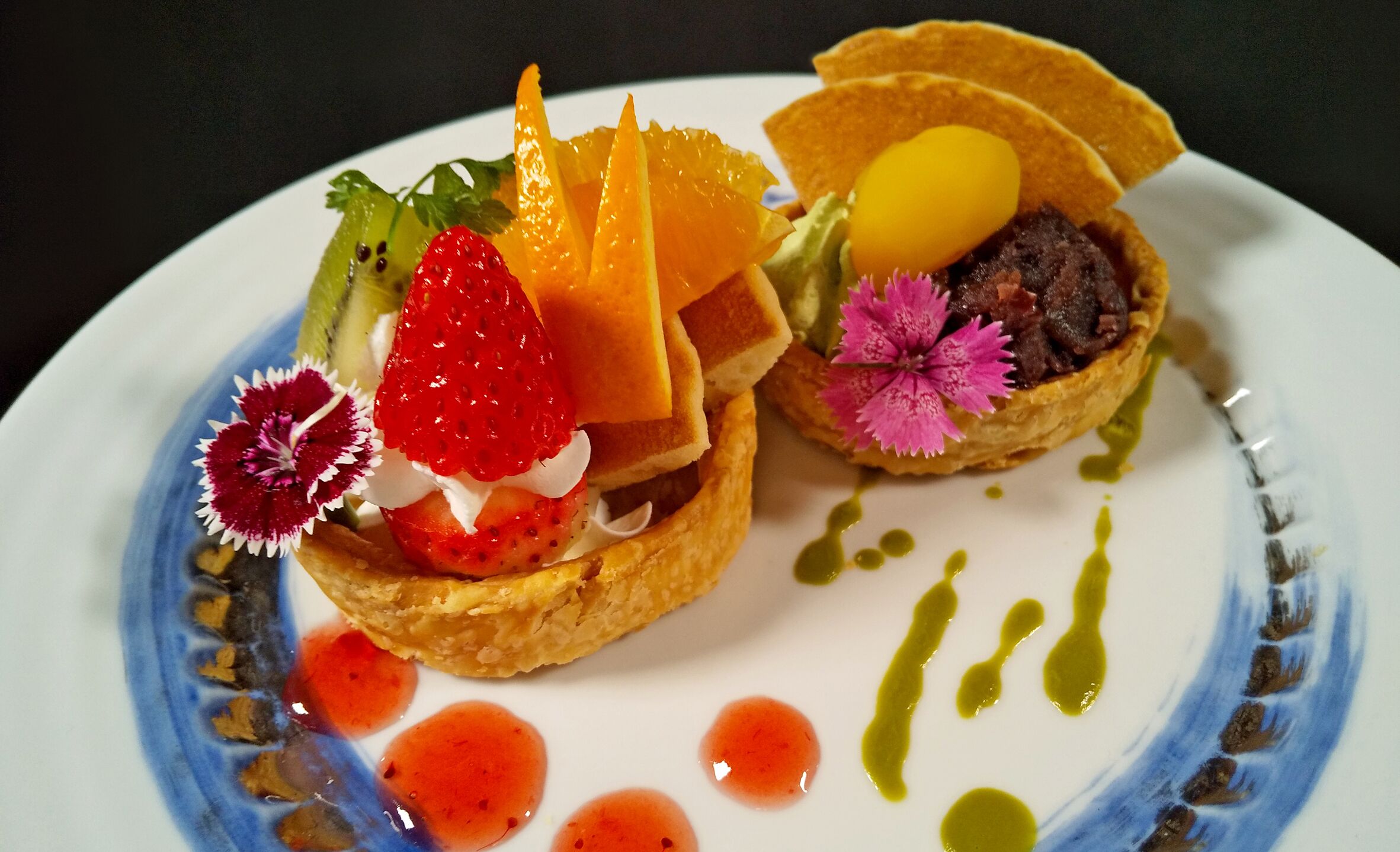 Un piatto con due piccoli dolci guarniti con fragole, formaggio e un fiore, irrorati da salse.