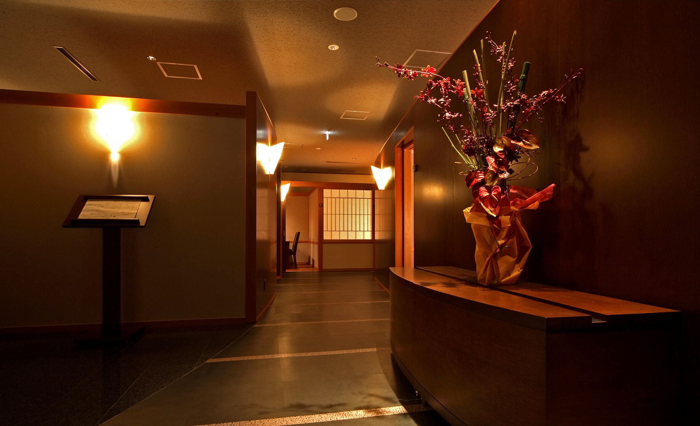 A hotel reception area with a reception desk, a wall-mounted information board, and a decorative floral arrangement.