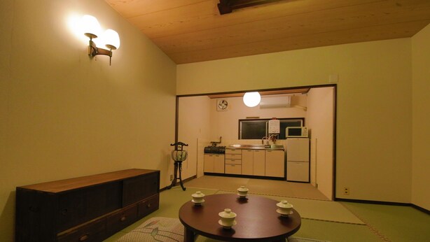 Una habitación japonesa tradicional con suelo de tatami, una mesa baja de madera con vajillas de té, un banco de madera y una cocina visible a través de un vano abierto.
