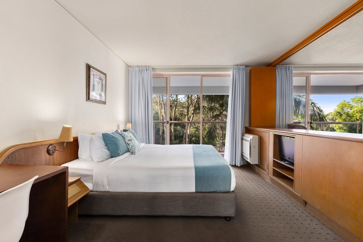 Une chambre d’hôtel équipée d’un lit, d’un bureau, d’une télévision et de grandes fenêtres avec des rideaux.