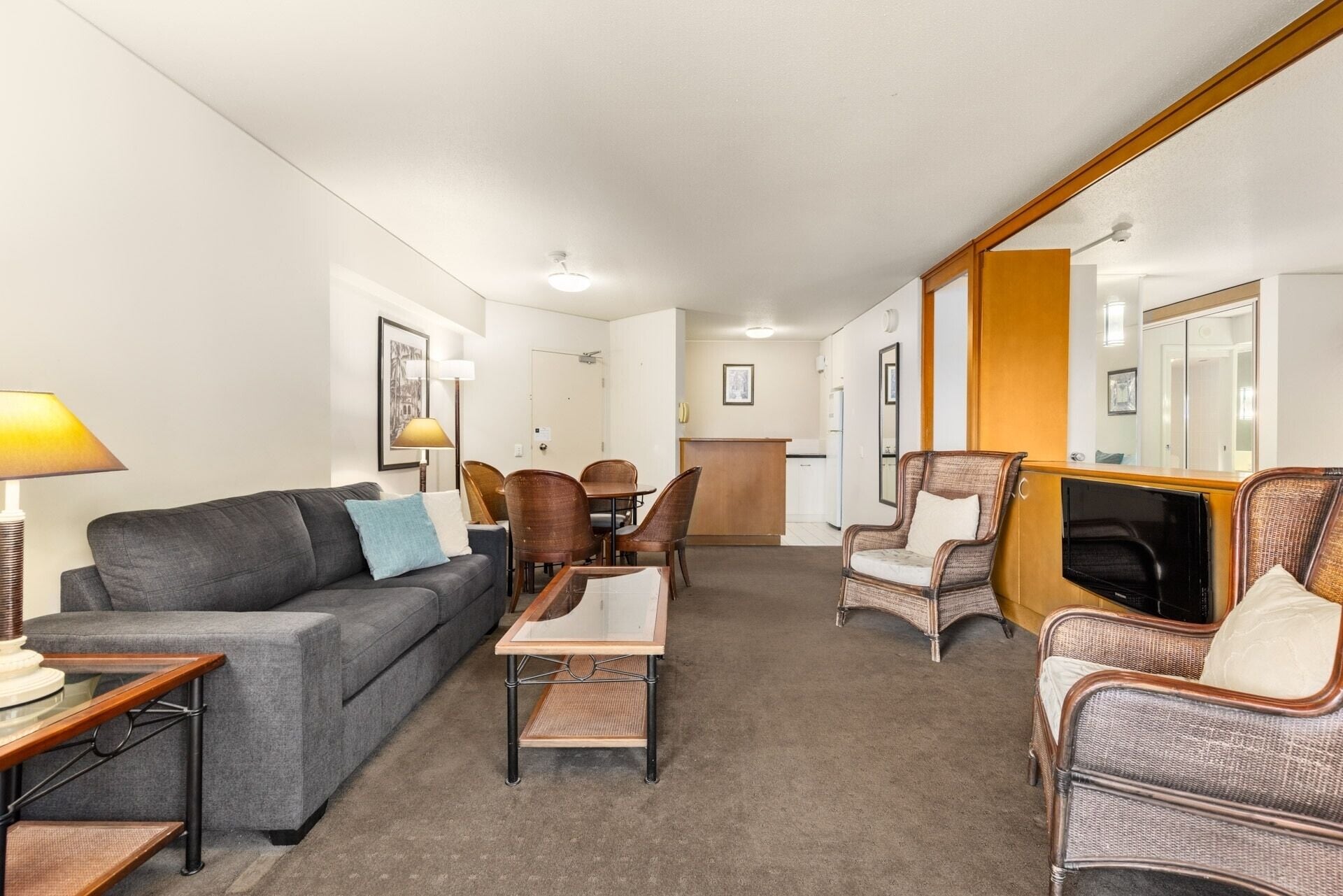 A modern hotel room with a grey sofa, a small table, wicker chairs, a TV, and a large mirror.