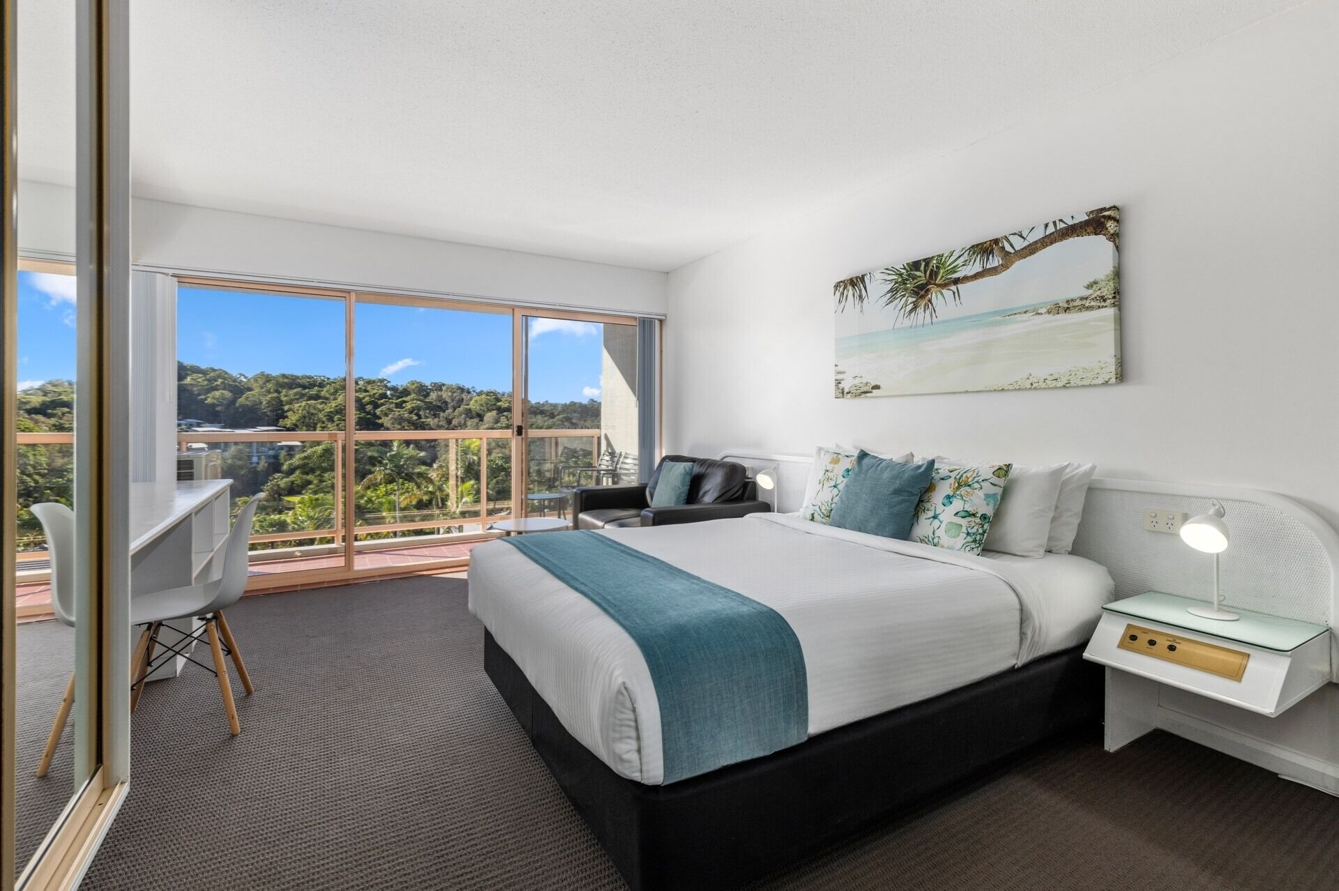 A modern bedroom with a large bed, a desk, and a chair. It features a balcony with a view of the city and greenery.