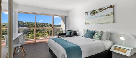 A modern bedroom with a large bed, a desk, and a chair. It features a balcony with a view of the city and greenery.