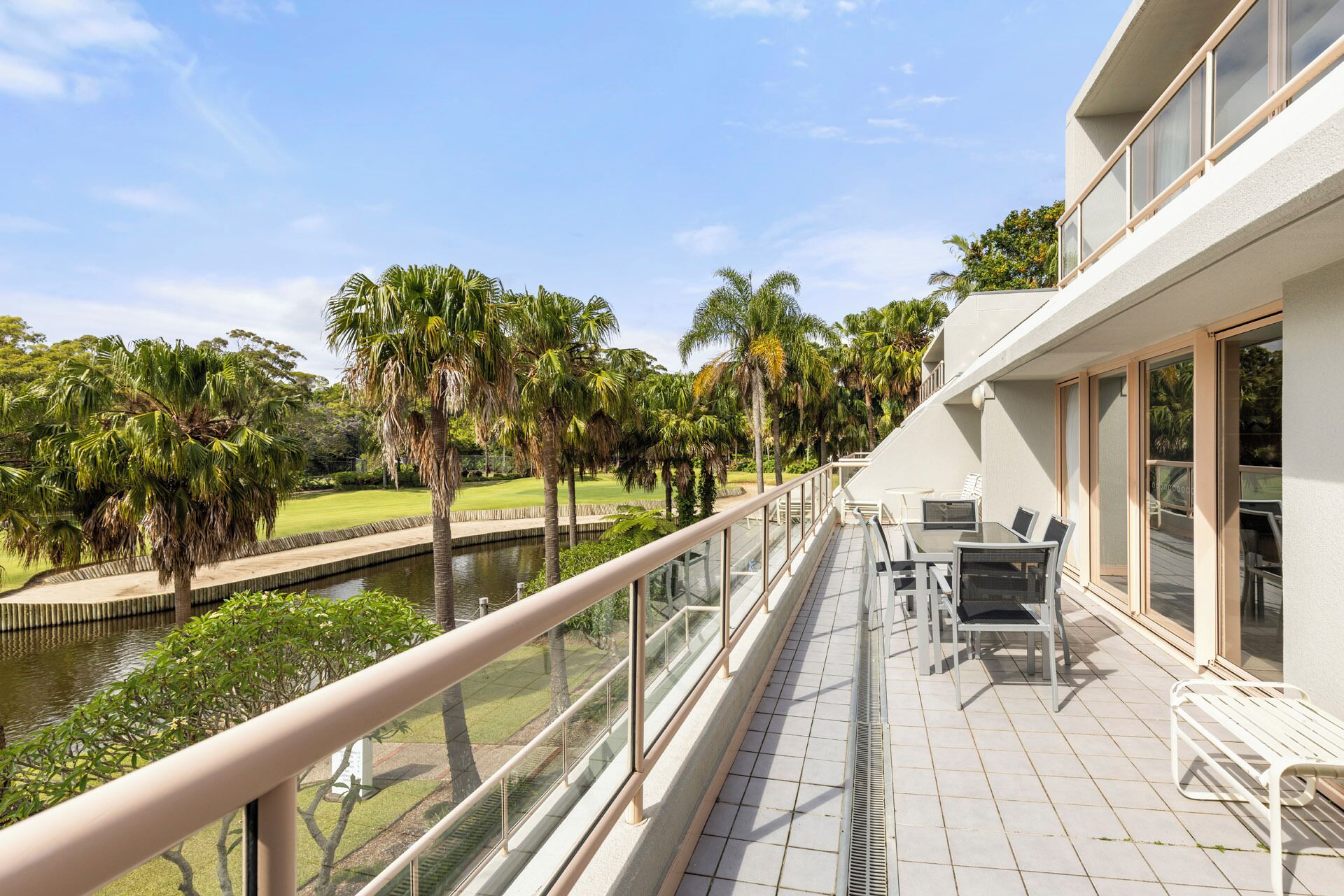 A balcony with a view of a water body and palm trees, a glass railing, and outdoor furniture.