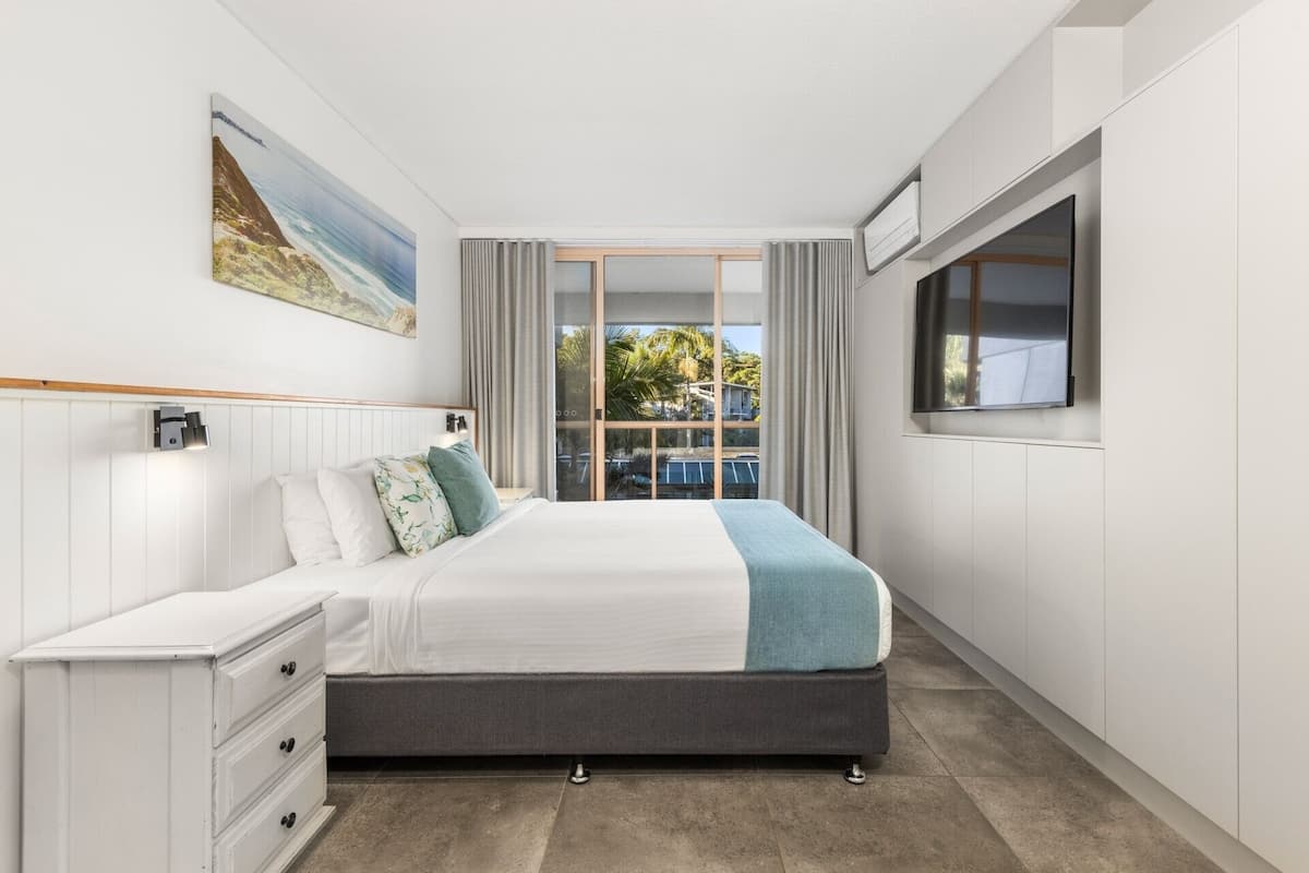 Une chambre à coucher avec un lit, une table de chevet, une commode, une fenêtre et un tableau accroché au mur.
