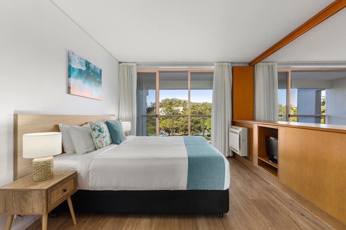 Une chambre d’hôtel avec un grand lit, une table de chevet avec une lampe, un téléviseur et une vue sur la verdure grâce à de grandes fenêtres.