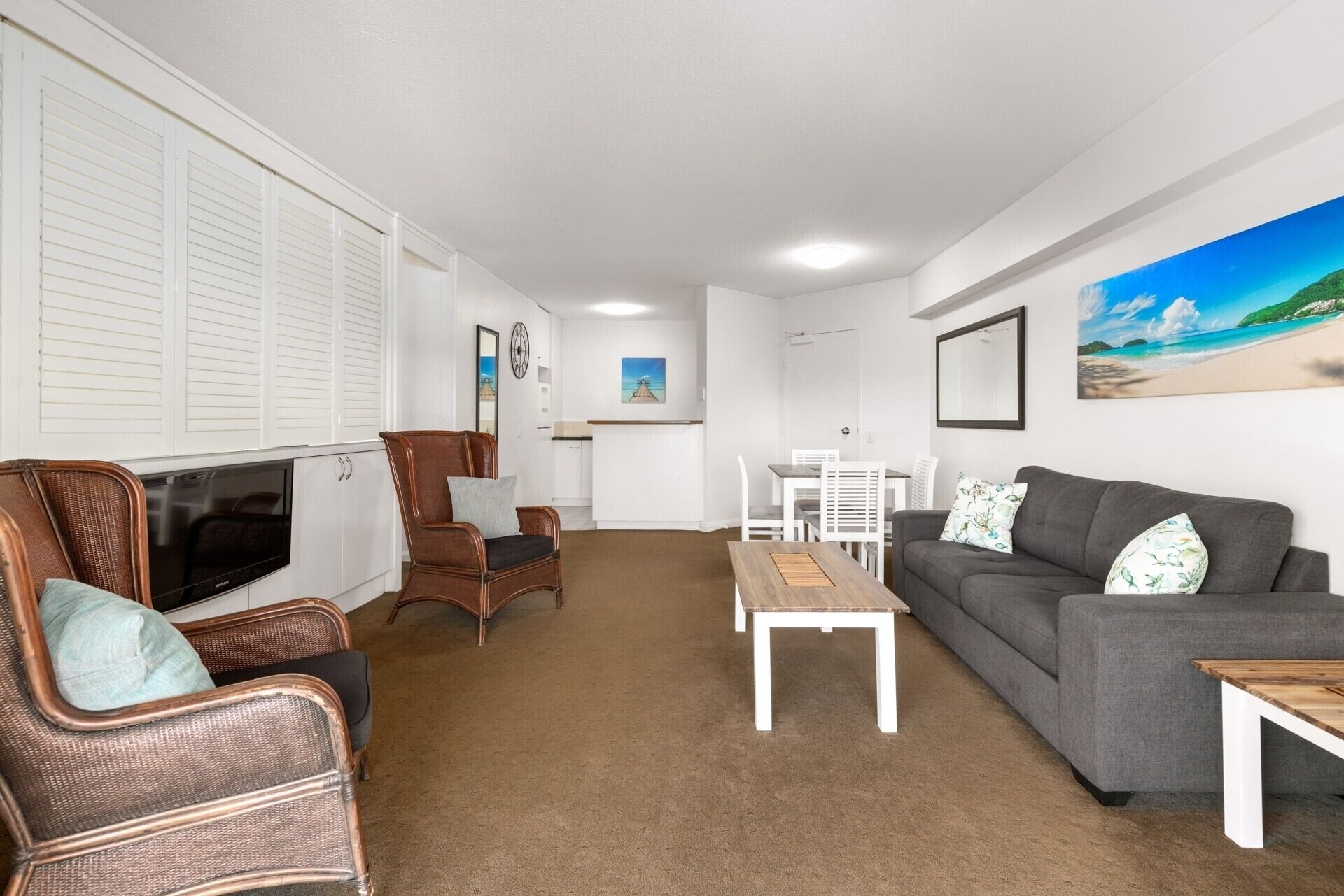 A living room with a grey sofa, wicker chairs, a wooden coffee table, a TV, and a dining area with a white table and chairs.
