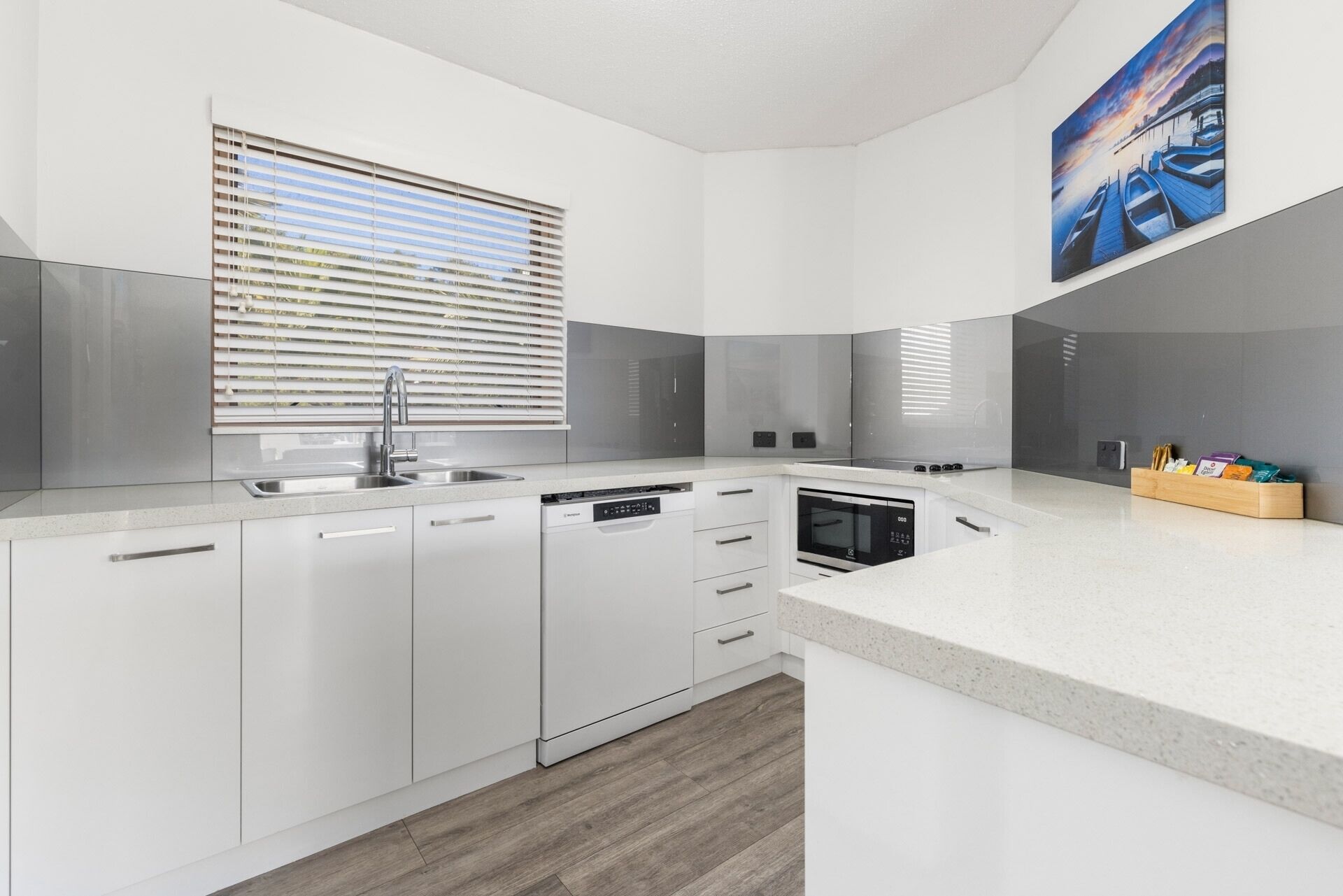 A modern kitchen with white cabinetry, a built-in dishwasher, and a large window with blinds.