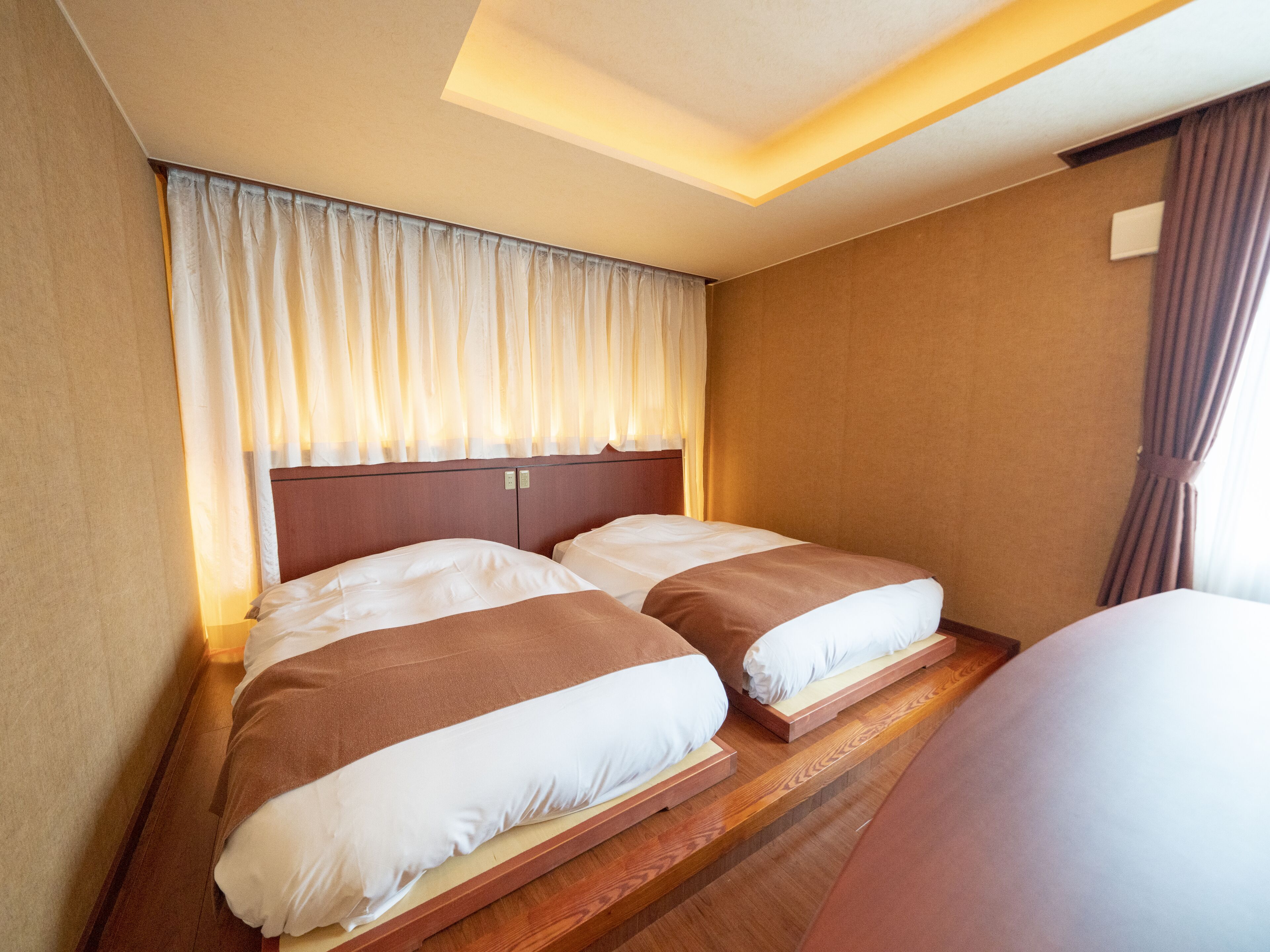 A hotel room with two beds, wooden flooring, a headboard, and a window with curtains.