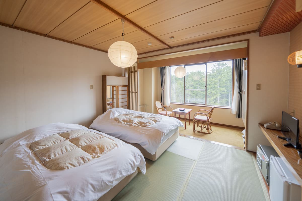A traditional Japanese-style bedroom with tatami flooring, two beds with white bedding, a desk with a chair, and a large window with a view of trees.