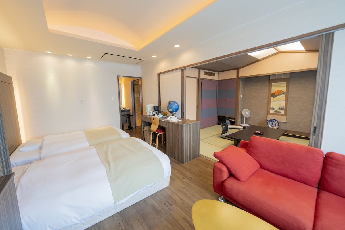 A hotel room with a bed, a desk, a chair, a red sofa, and a small table with a globe and a lamp.