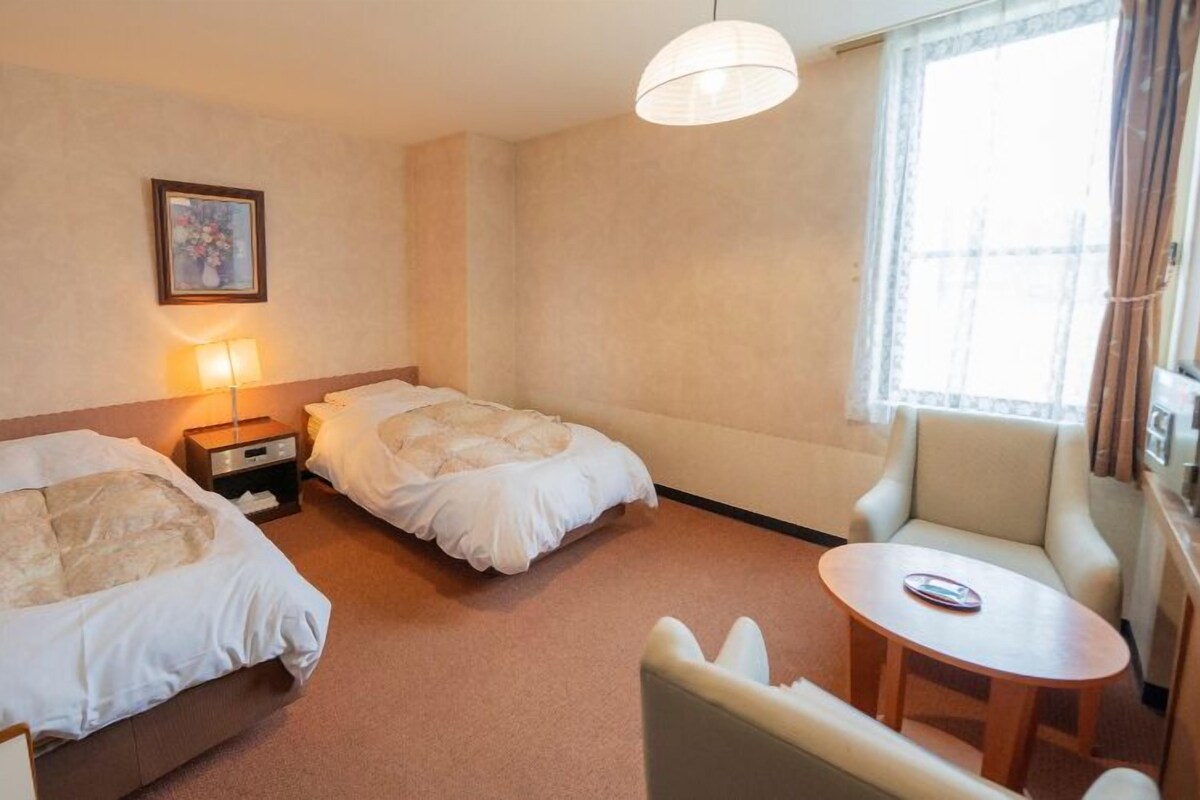 A hotel room with two beds, a small table, a chair, a lamp, and a picture on the wall.