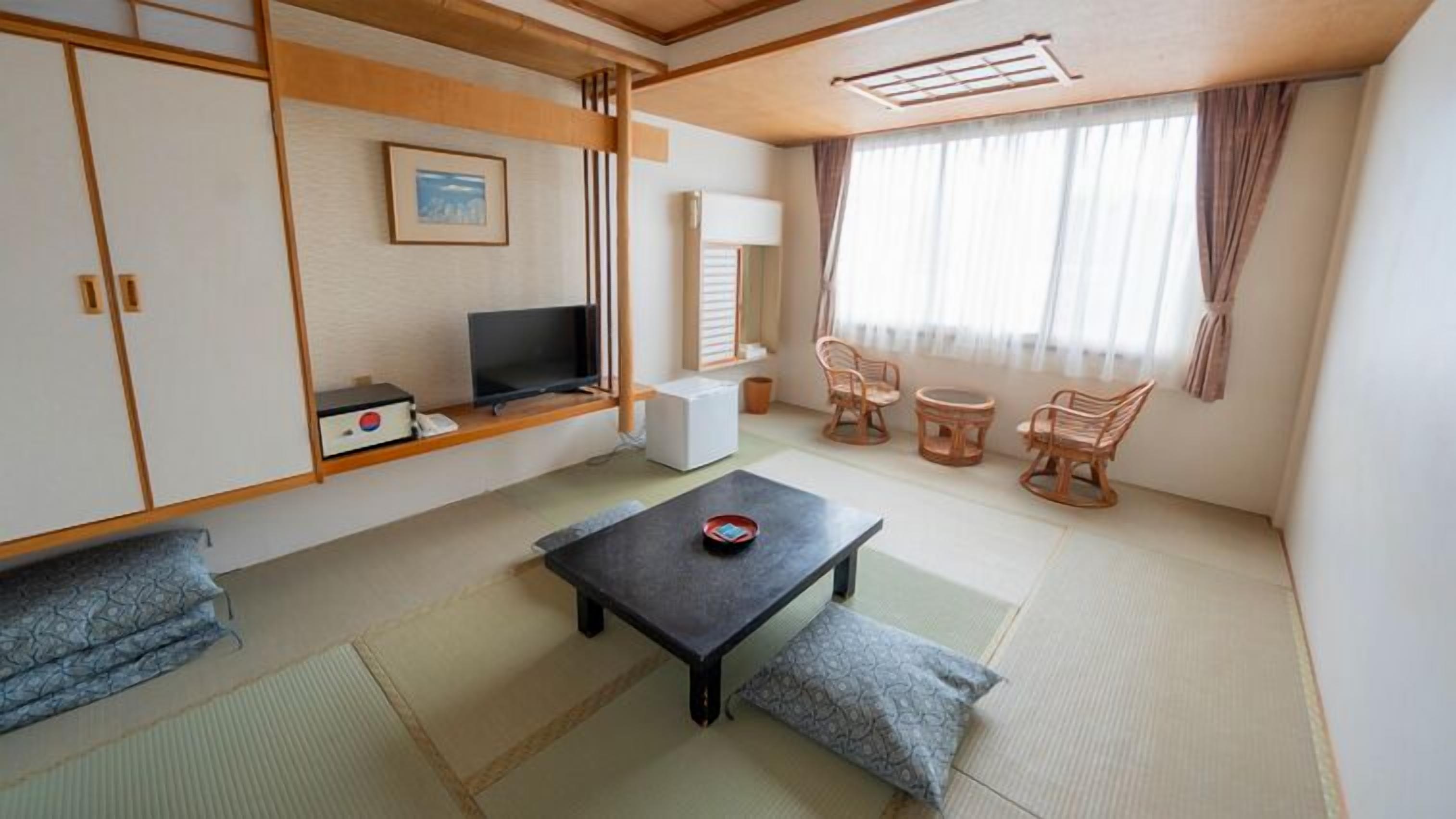 A traditional Japanese-style room with tatami flooring, a low table with cushions, a television, and a window with curtains.