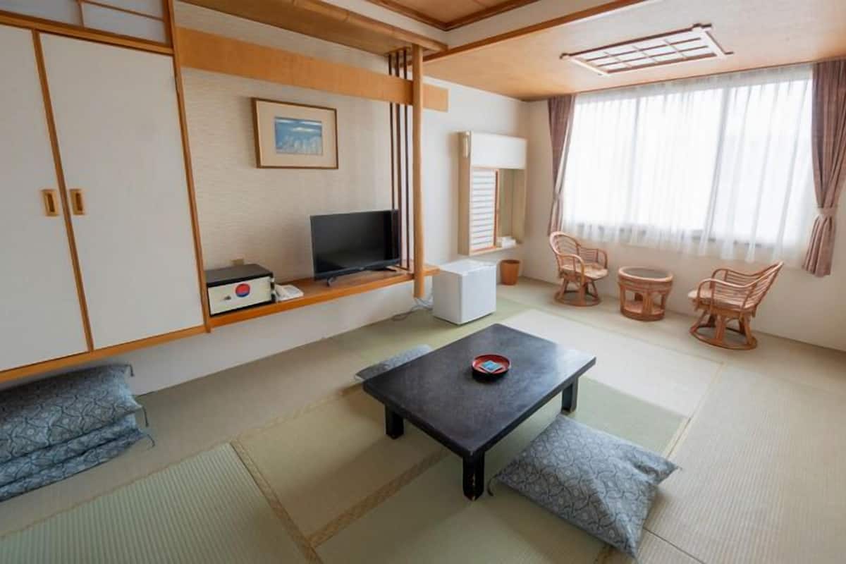 A traditional Japanese-style room with tatami flooring, a low table with cushions, a television, and a window with curtains.