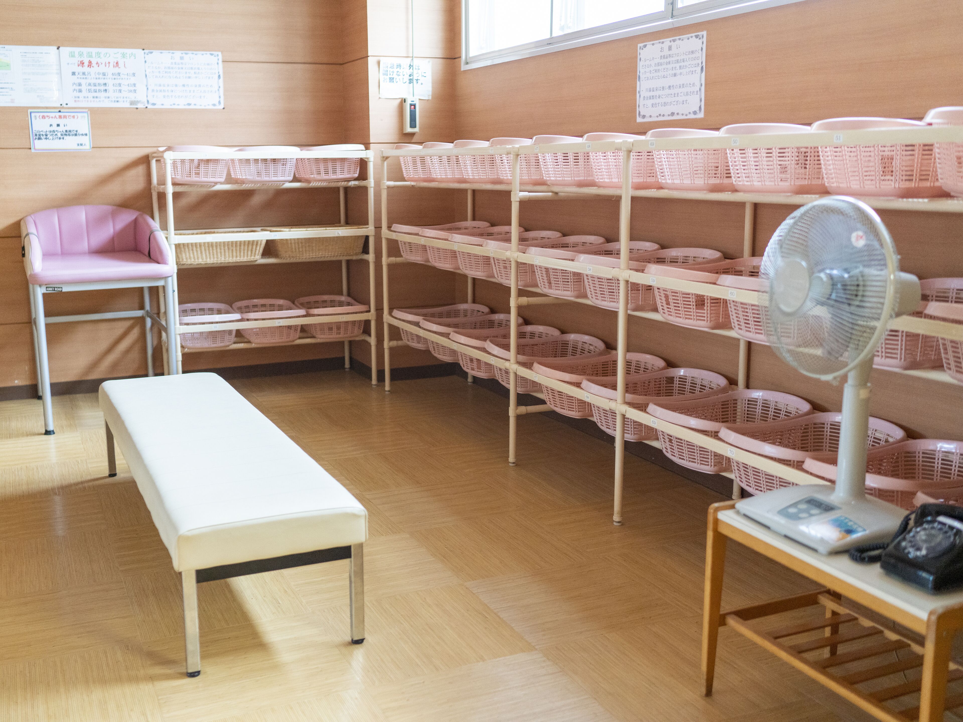 A room with wooden flooring, a white bench, pink baskets on shelves, a pink chair, and a fan on a table.