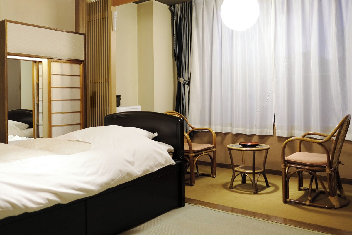 A hotel room with a bed, a chair, a small table, a mirror, and a window with curtains.